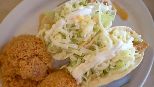 Top view of two chicken tacos filled with shredded lettuce, cheese, and sauce, served with a side of Mexican rice on a white plate.