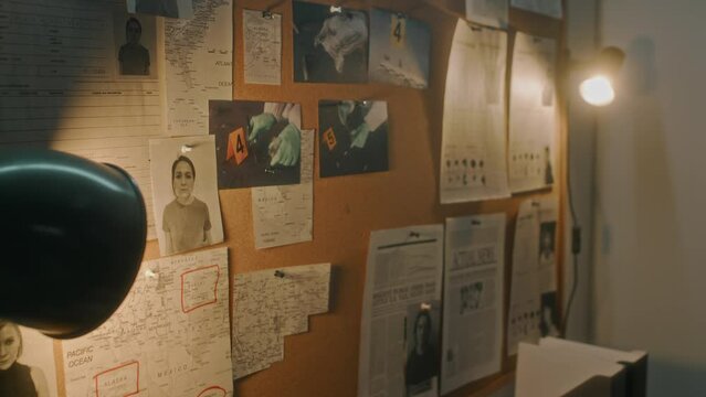 Close-up tilt down shot of pin board on wall in detectives office of police department, with photos of male, female crime suspects, murder scene images, maps, schemes, illuminated by lamps