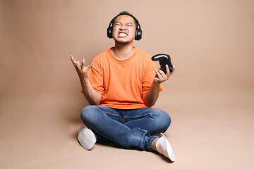 Upset Asian guy sitting on the floor losing video games isolated on beige background
