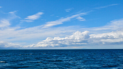 View of the blue sea and white clouds above the horizon. View of small waves on the sea surface and thick white cumulus clouds in the blue sky above the horizon.