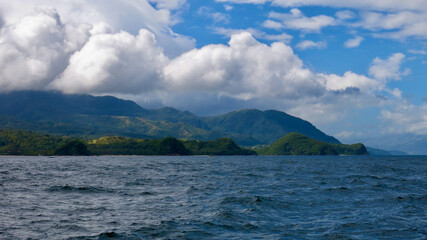 View from the sea to a tropical island and cumulus clouds. Coastal view of a hilly tropical island and cumulus clouds over a mountain range. Tropical landscape.