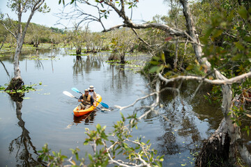 Holiday travel activities. Happy asian couple man and woman rowing a canoe or kayak in mangrove forests. Young traveler with kayak at botanical garden tropical mangrove forest in a national park.
