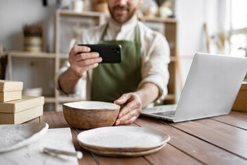 Positive manager of decorative clay store takes photo on phone for social media. Mature man, potter, engaged in making home-made dishes and updating website portfolio.