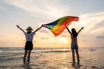 Young couple asian lesbian with pride movement LGBT holding rainbow flag for freedom. Demonstrate rights LGBTQ celebration pride Month lesbian Pride Symbol. Walking on the sand sea beach with sunset