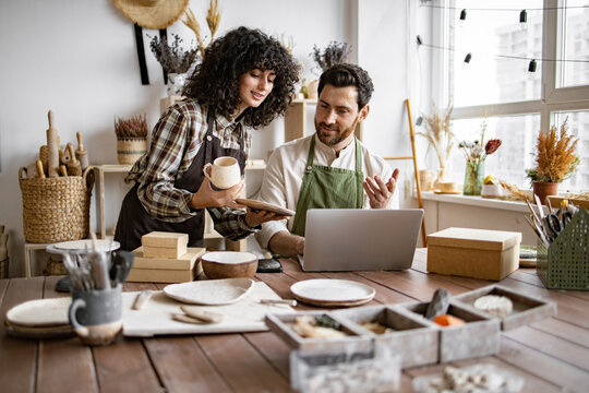 Caucasian couple of co-workers work on small pottery production business. Mature man and young curly-haired woman in aprons use laptop to take inventory of products on store website. - Powered by Adobe
