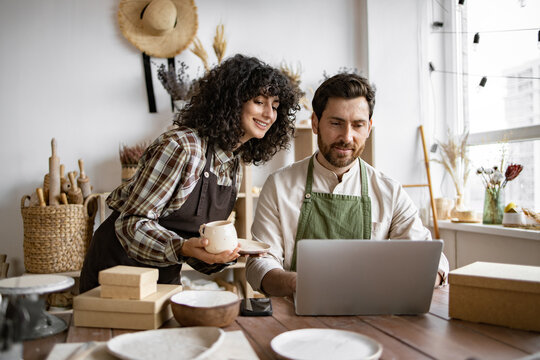 Caucasian couple of co-workers work on small pottery production business. Mature man and young curly-haired woman in aprons use laptop to take inventory of products on store website.