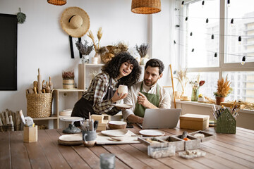 Portrait of mature man and young curly woman creating online catalogue of available goods at decor store. Two workers holding original ceramic products, using laptop during working at shop. - Powered by Adobe
