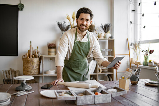 Concept of small business, handmade products and online sales on Internet. Bearded adult man in apron and white shirt viewing an online course on pottery and making ceramic dishes.