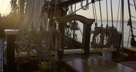 1700s sailing ship underway at sunrise. Static medium shot. Sun burst causing lens flare across the ship's bell and pin rails.