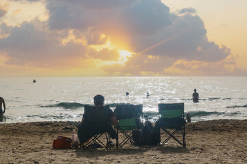 People relaxing on the beach at sun sets. Man sit in chair on a beach, watching the sunrise over the sea. Clouds adorn the sky and sun rays dance on the water surface. Beachgoers relax facing the sea
