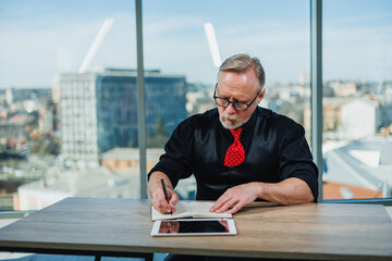A gray-haired man in a black shirt sits at a desk in the office and works with a tablet. Modern...