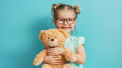 Cute little girl wearing glasses hugging teddy bear on blue background. Happy caucasian child of 5-6 years looking at camera and smiling. Preparation for school, courses, children's clinic