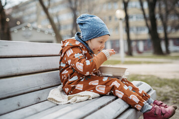 Cute toddler girl eating lunch in park, sitting on bench. Picnic in park during cold spring day.