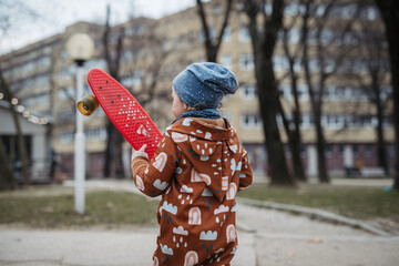 Cute toddler girl playing outdoor in playground with skateboard. Girl in softshell bodysuit...
