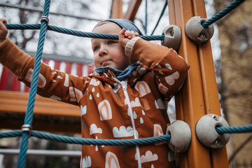 Cute toddler girl playing outdoor in playground, climbing jungle gym. Girl in softshell bodysuit...