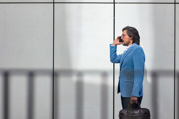 Portrait of mature businessman making phone call, standing on city street, in front of office...