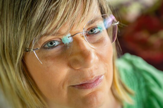 Close-up portrait of a woman wearing glasses, highlighting her blue eyes and facial features. She has short blonde hair and wears a green top. The image is detailed, capturing the subtle expressions - Powered by Adobe