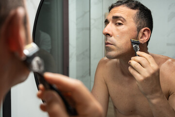 Young man meticulously grooms sideburns using razor's edge in bathroom mirror, emphasizing male...