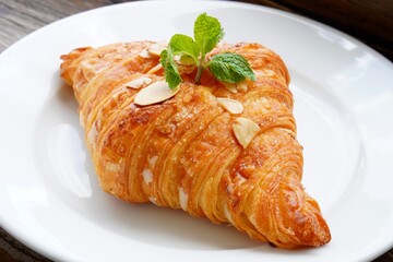 A butter croissant delicately dusted with sugar and sliced almonds, topped with a fresh mint leaf, elegantly presented on a white plate.