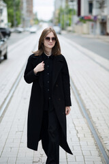 Fototapeta premium Attractive Caucasian young woman in sunglasses and black coat, walking along city street on autumn day. Model in stylish clothes. Outdoors portrait. Lifestyle, fashion Concept.