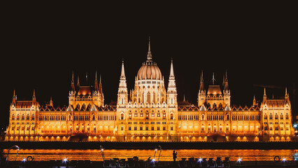 Fototapeta premium Panoramic background orange night image of Budapest parliament from the front side with boat and tourist taking a photo of the city. Travel in hungary and historical sites.