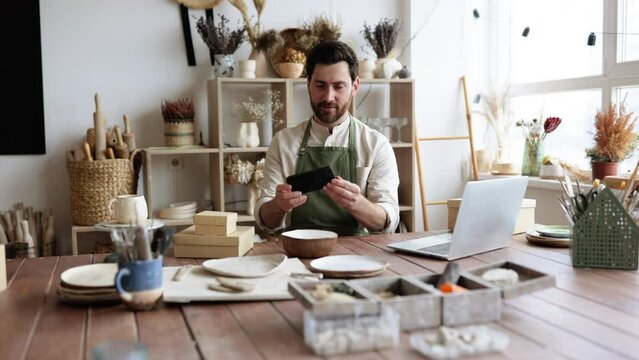 Charismatic Caucasian man seller in souvenir shop takes pictures of goods at phone for posting on social networks. Successful guy store employee with ceramic tableware shoots photos on smartphone.
