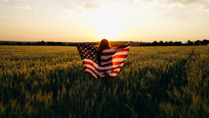 Young  woman with American flag in a wheat field at sunset celebrate Independence day. 4th of July....