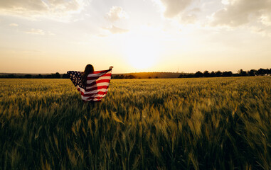 Young  woman with American flag in a wheat field at sunset celebrate Independence day. 4th of July....