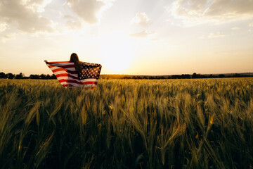 Young  woman with American flag in a wheat field at sunset celebrate Independence day. 4th of July....