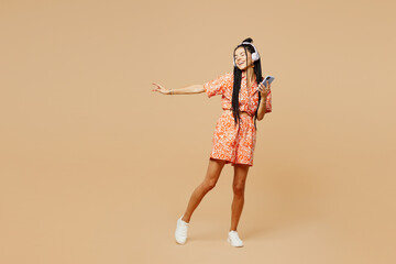 Full body happy young Latin woman she wear orange casual clothes listen to music in headphones use mobile cell phone isolated on plain pastel light beige background studio portrait. Lifestyle concept
