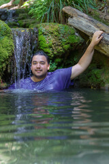 attractive young dark-haired boy cooling off under the waterfall of a river wearing a blue T-shirt