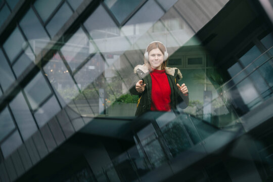 Smiling woman wearing wireless headphones and unlocking car with remote control key seen through glass window