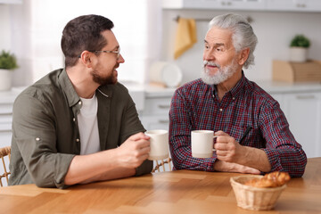 Happy son and his dad with cups talking in kitchen