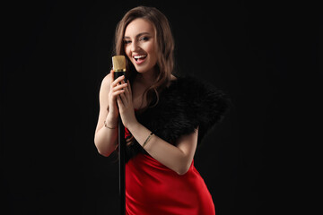 Beautiful young woman in stylish dress with microphone singing on black background