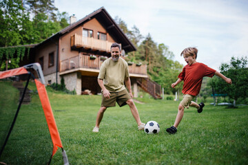 Dad having fun with young son, playing football, running and kicking the ball. Playing on a lawn in...