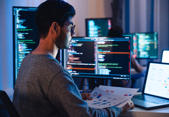 Asian software developers working on multiple screens displaying code and application diagrams at night in modern office