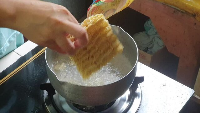 POV hands cooking instant noodle soup at home. Asian female hand putting dry noodles into boiling water in a pan at home and stirring. Steaming food on gas stove.
