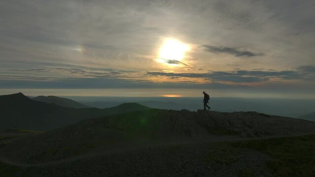 Mountain walker approaching summit silhouetted by golden hour sun. Wide shot. Grisedale Pike, Lake District, Cumbria, England, UK