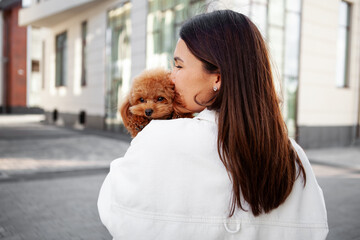 Woman gently hugs and kisses small dog outdoors in an urban setting. Affection and bond between the...