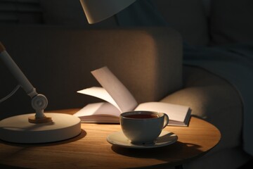 Stylish lamp, book and cup of tea on side table near sofa indoors