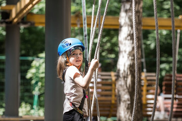 Summer. A small child is climbing a rope bridge in a rope park. A girl is having fun in an adventure park. A female kid on a climbing wall. Compliance with safety regulations.