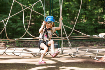 Summer. A small child is climbing a rope bridge in a rope park. A girl is having fun in an adventure park. A female kid on a climbing wall. Compliance with safety regulations.