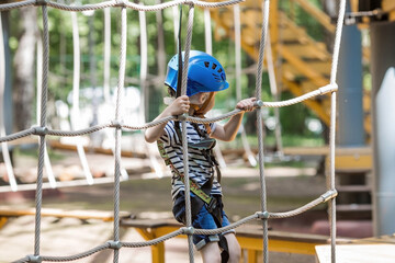 Summer. A small child climbs in a rope park on a rope bridge. A boy is having fun in an Adventure Park. A male baby on a climbing frame. Compliance with safety techniques.