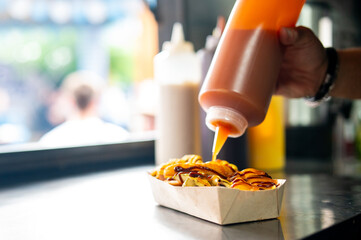 hand squeezes sauce onto a hot dog with toppings in a cardboard tray, suggesting a street food setting