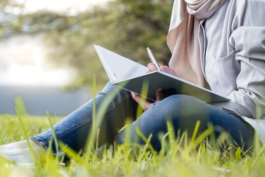 Young Asian muslim woman writing in a notebook while sitting on grass in a sunny morning during summer at the park