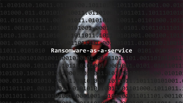 Ransomware-as-a-service cyber attack text in foreground screen, anonymous hacker hidden with hoodie in the blurred background. Vulnerability text in binary system code on editor program.