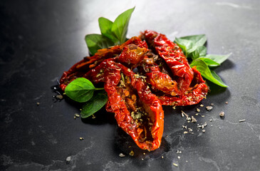 Sun dried tomatoes with aromatic herbs, oregano, marjoram leaf. Over black stone background, close up. Italian cuisine, healthy vegan food, Mediterranean diet. Dry tomato