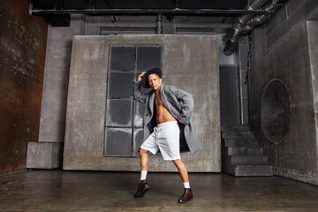 Male dancer, wearing gray coat, white shorts and black hat, performs in studio with grey wall in...