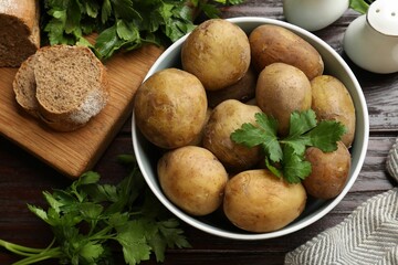 Boiled young potatoes in bowl, parsley and bread on wooden table, top view