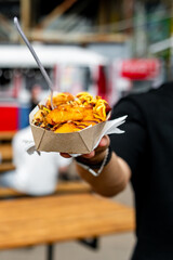Close-up of hand holding street food paper tray with grilled items and toppings outdoors.